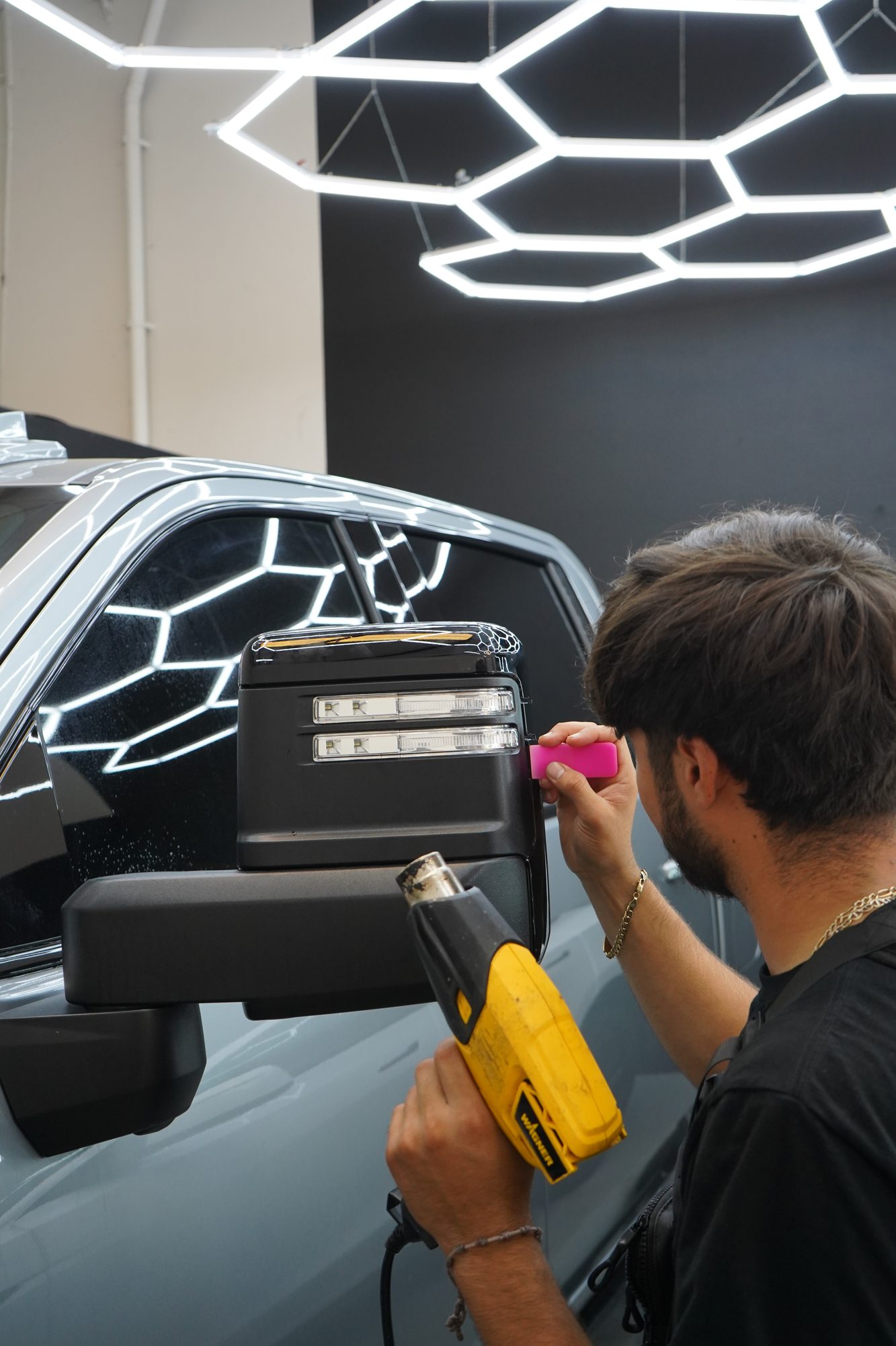 An installer using a heat gun to form paint protection film around a GMC Sierra's tow mirror, under the hex lights